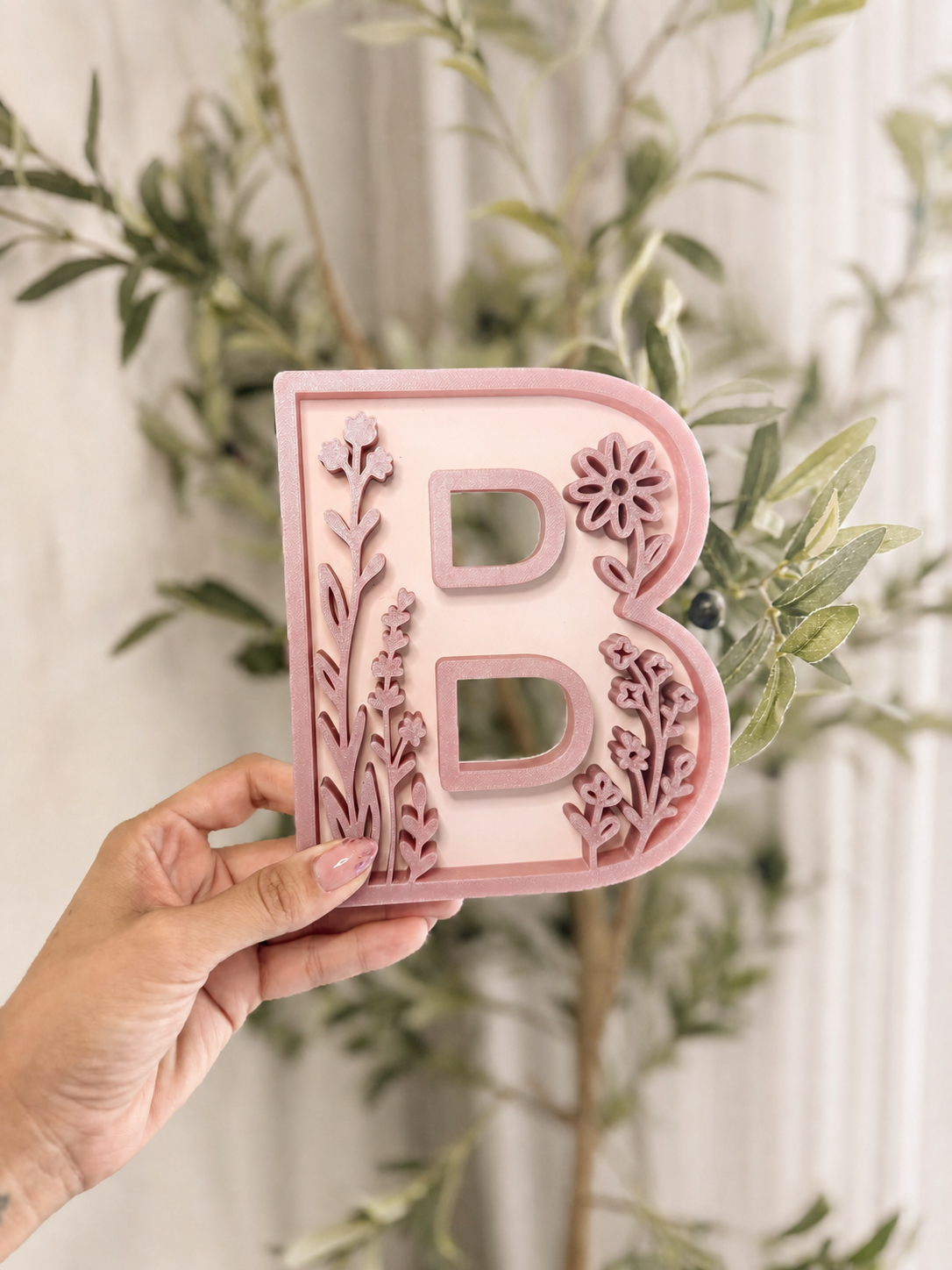 Blush pink floral letter B nursery decor held against a softly lit leafy background